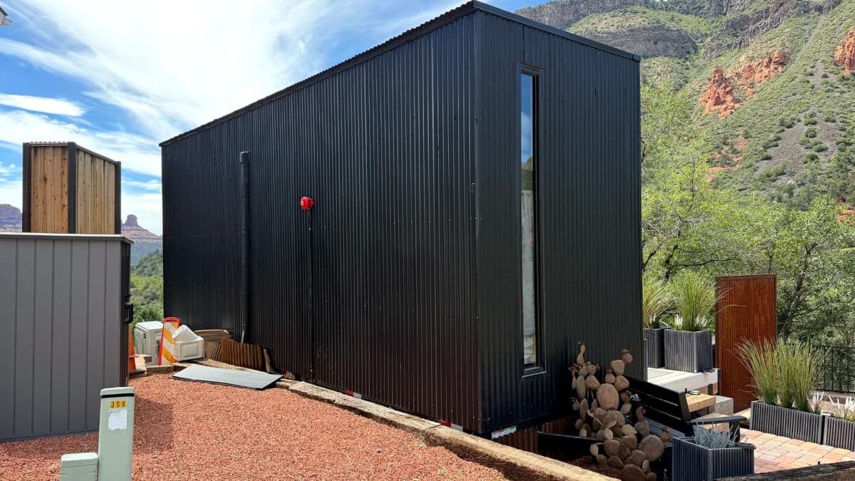Black corrugated container home with tall narrow window set against a red rock mountain landscape