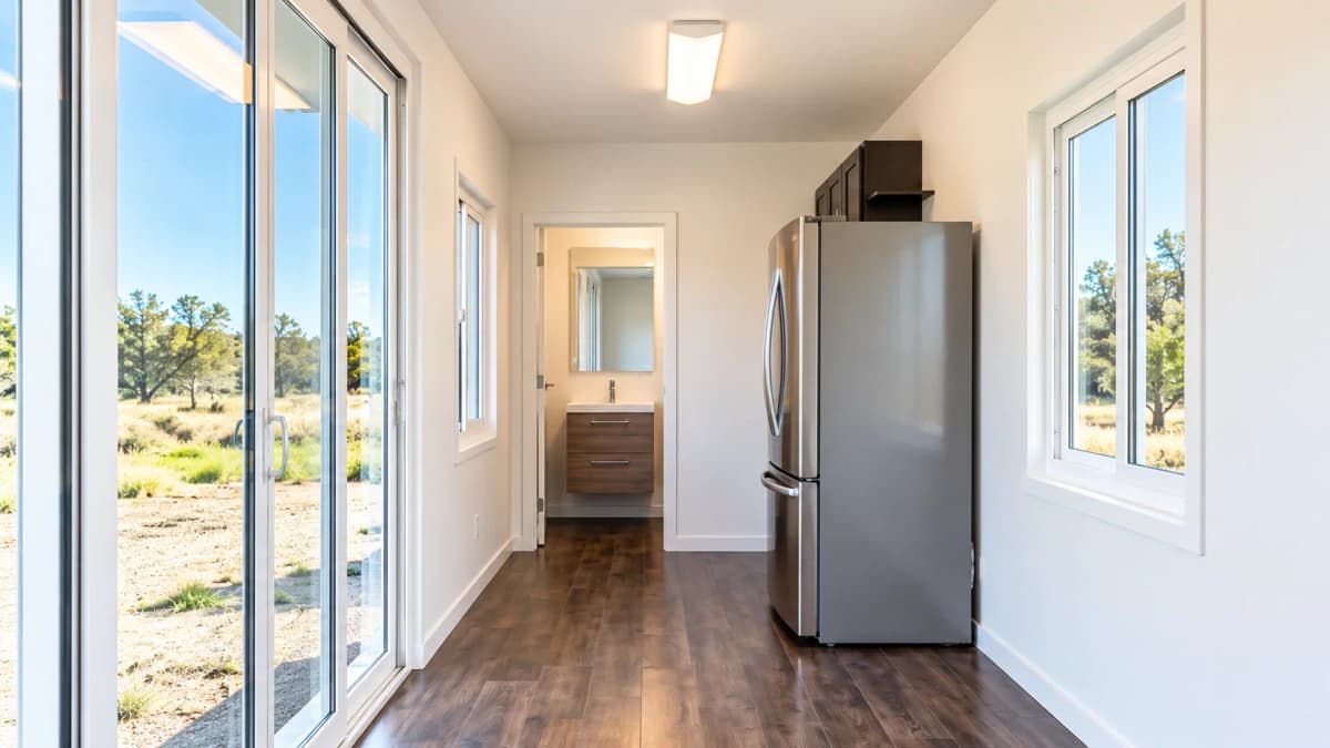 Container studio interior with stainless steel refrigerator, wood-look flooring, large glass sliding doors, windows, and finished bathroom beyond