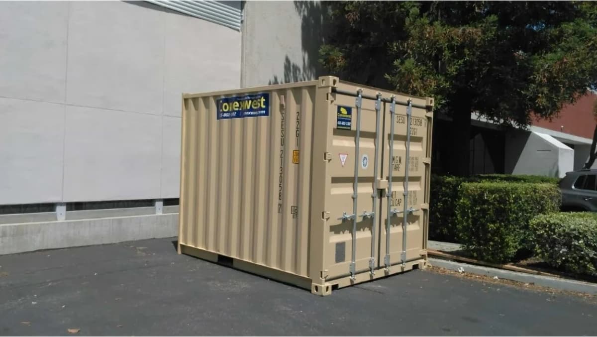 A beige shipping container in a lot in front of a building with hedges and trees behind it