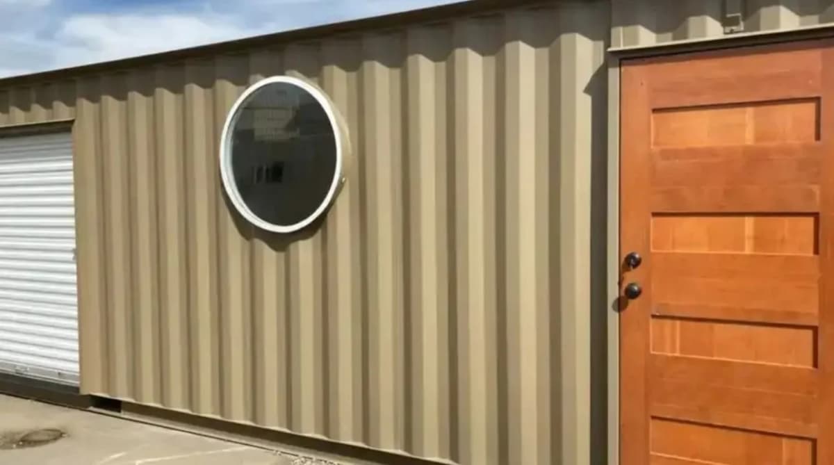 A beige shipping container with a round window, wooden door, and pull-up garage door.