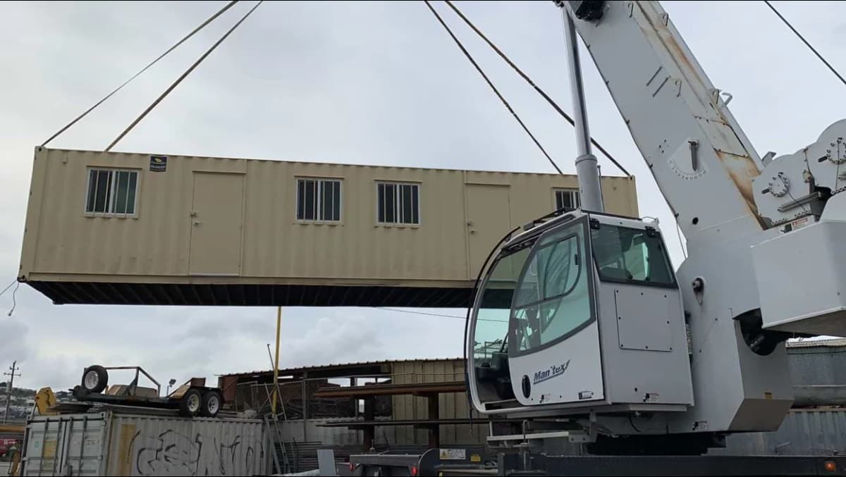 A crane hoisting a beige Conexwest shipping container converted into an office unit, featuring multiple windows and doors, at an industrial site.