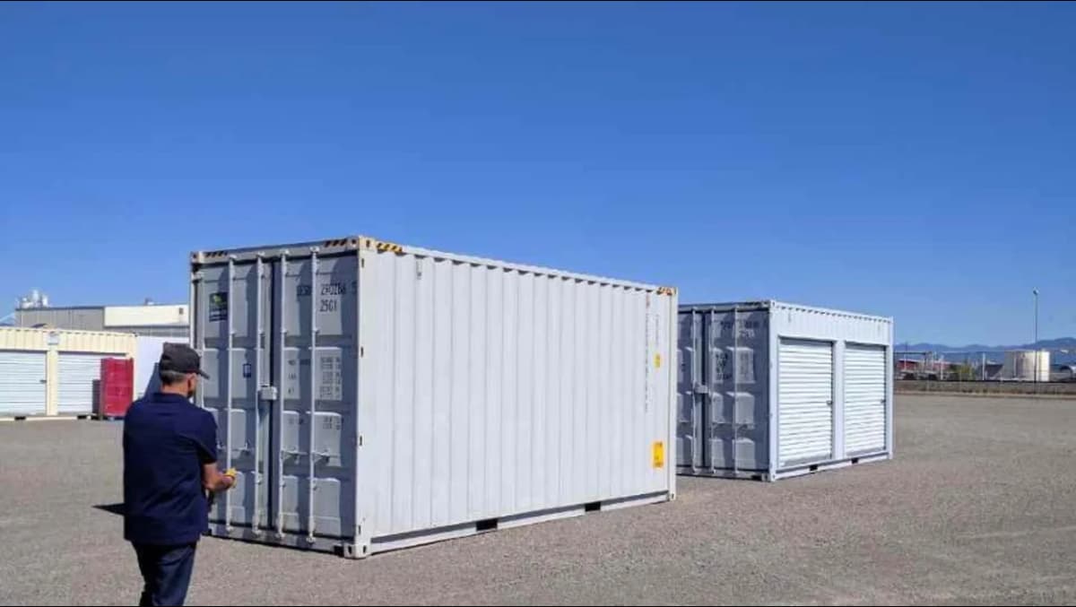 A man standing near two Conexwest industrial shipping containers placed in a large open lot.