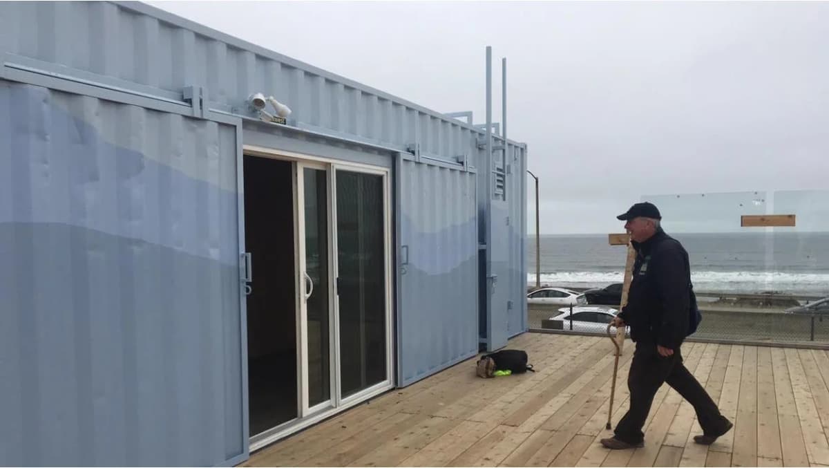 A man with a walking stick approaches a modern shipping container home with sliding glass doors and a wooden deck overlooking the ocean