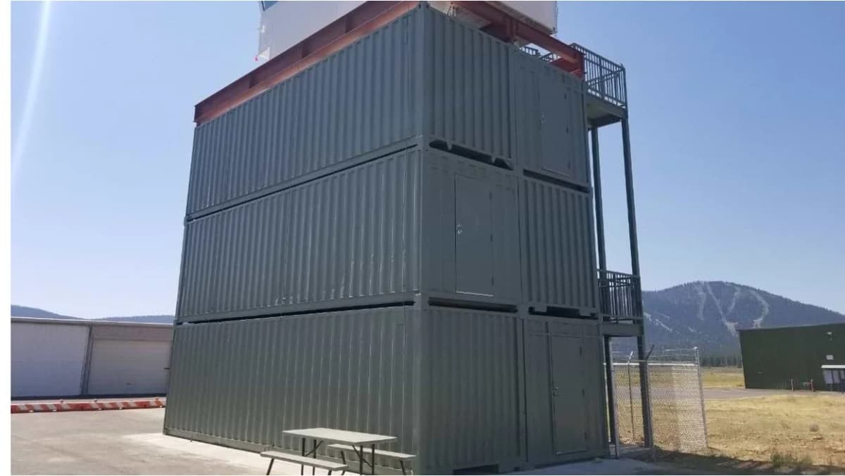 A tall structure made from stacked green shipping containers with staircases and balconies with mountains in the background