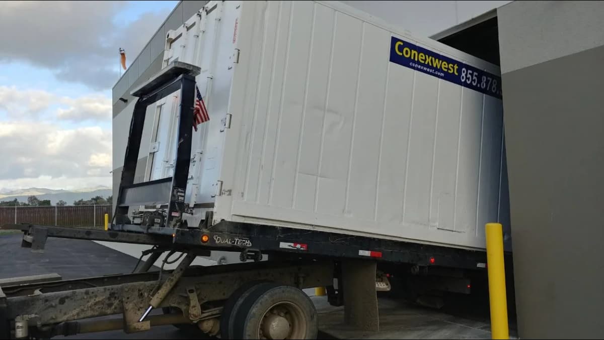 A white Conexwest container being delivered on a tilt-bed truck, partially backed into a building loading bay.