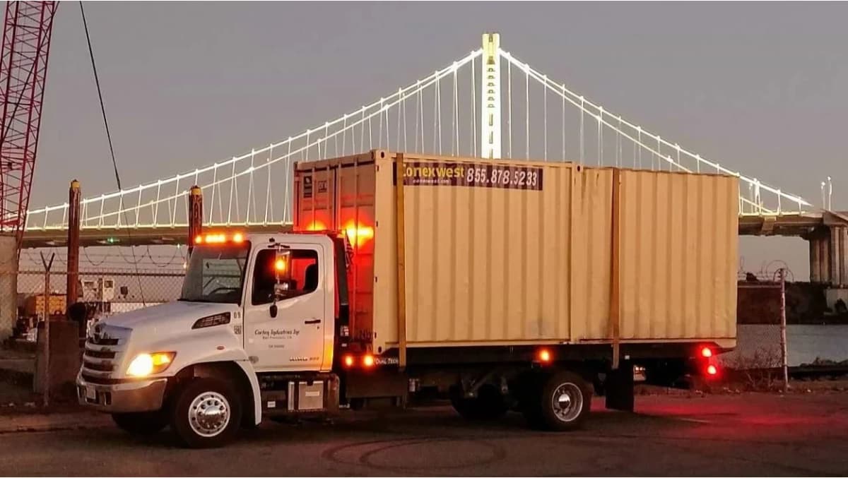 A white flatbed truck transporting a beige shipping container at dusk.