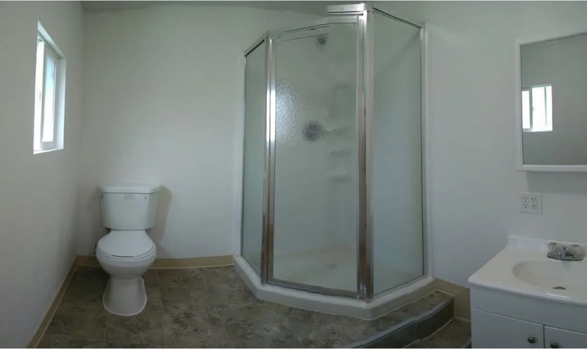 Bathroom inside a shipping container dorm with a corner shower, toilet, and vanity sink.