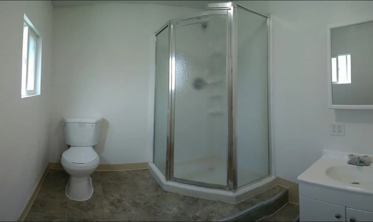 Compact bathroom with a corner glass shower, white toilet, and small vanity sink, featuring neutral-colored flooring and minimal decor.