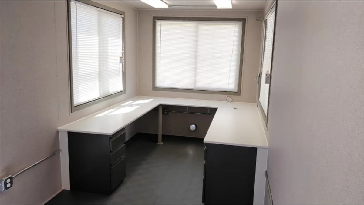 Compact office setup inside a container unit featuring an L-shaped white desk with black drawers, two windows with blinds, and overhead fluorescent lighting