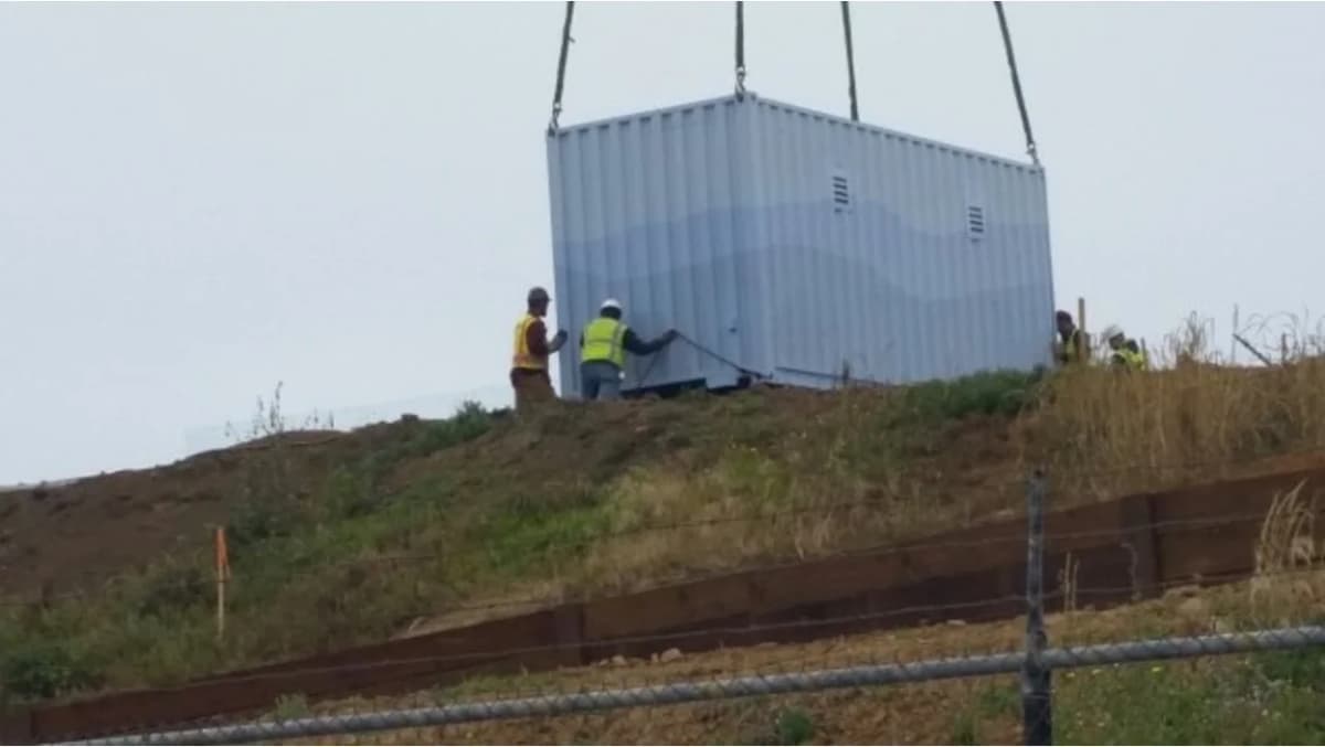 Construction workers in safety gear supervising the crane lifting of a white shipping container.