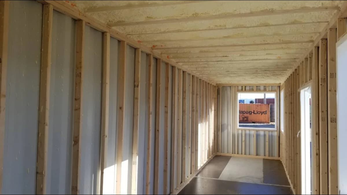 Interior construction view of a shipping container showing exposed corrugated steel walls, wooden wall studs, and ceiling beams.