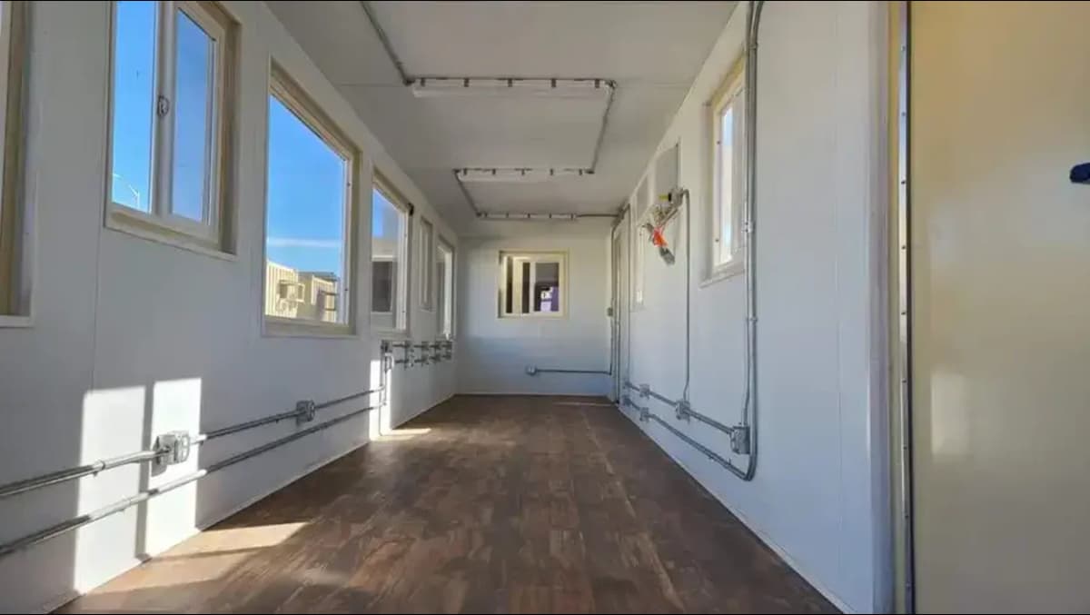 Interior corridor of a white container conversion with wood laminate flooring, multiple windows providing natural light, exposed wall fixtures, and track lighting on the ceiling.