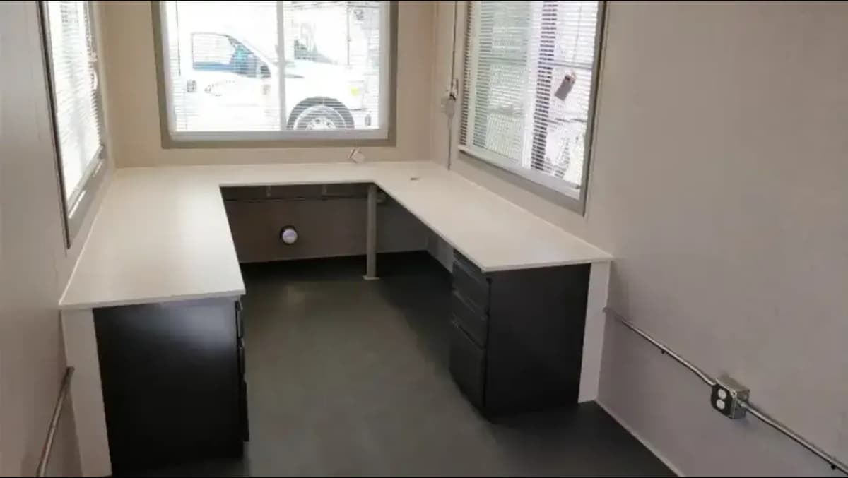 Interior of a compact container office with L-shaped white desks, black drawer units, and large windows on two sides.