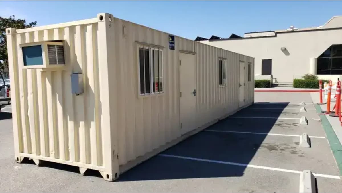 Long beige shipping container converted into an office unit with barred windows, dual entry doors, and a wall-mounted air conditioning unit, placed in a parking lot.
