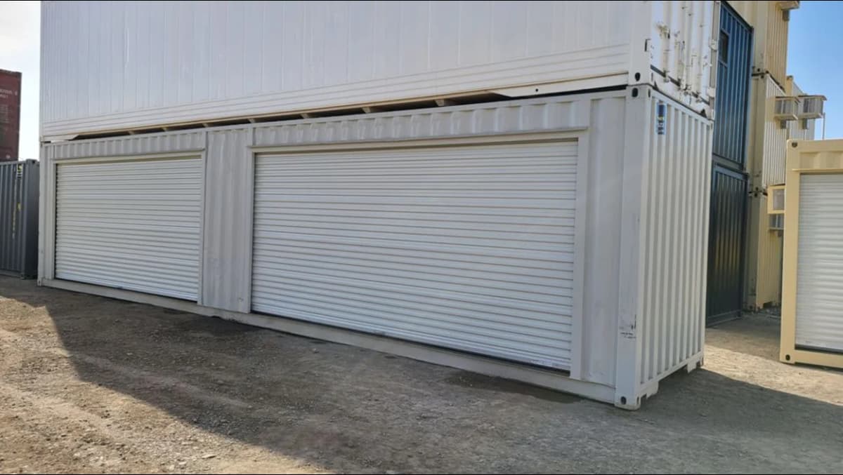 Stacked white shipping containers, with the lower container featuring two large white roll-up doors.