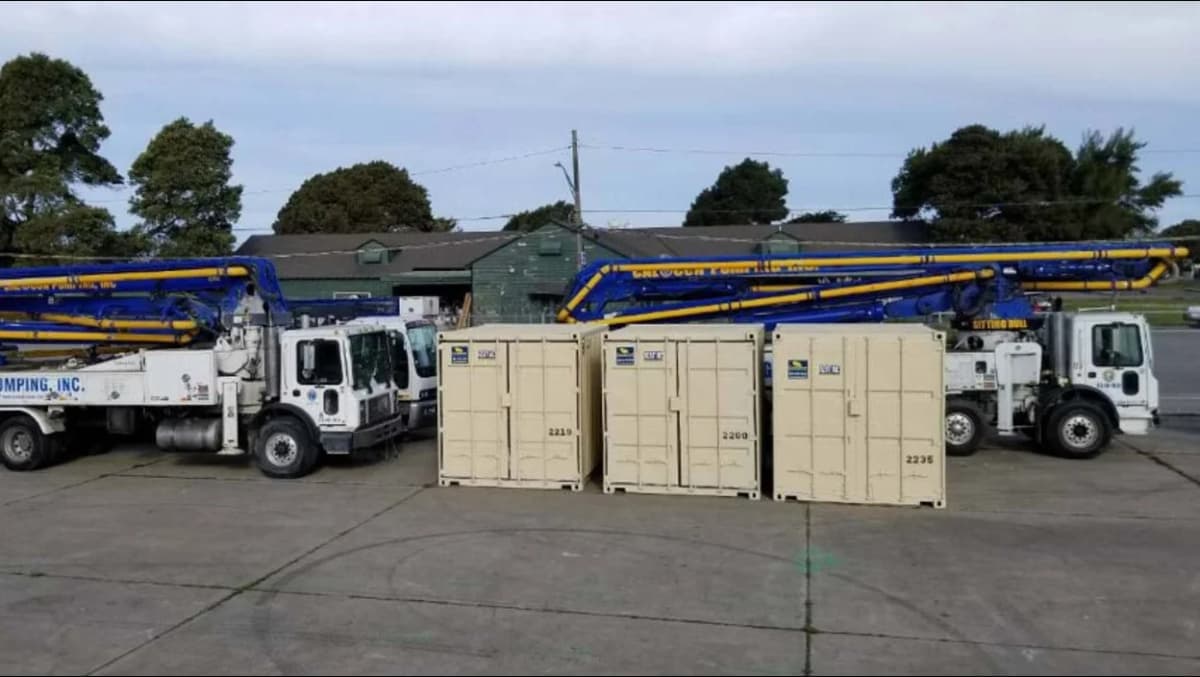 Three beige Conexwest storage containers are lined up in front of two large concrete pumping trucks with blue and yellow booms extended across the top.
