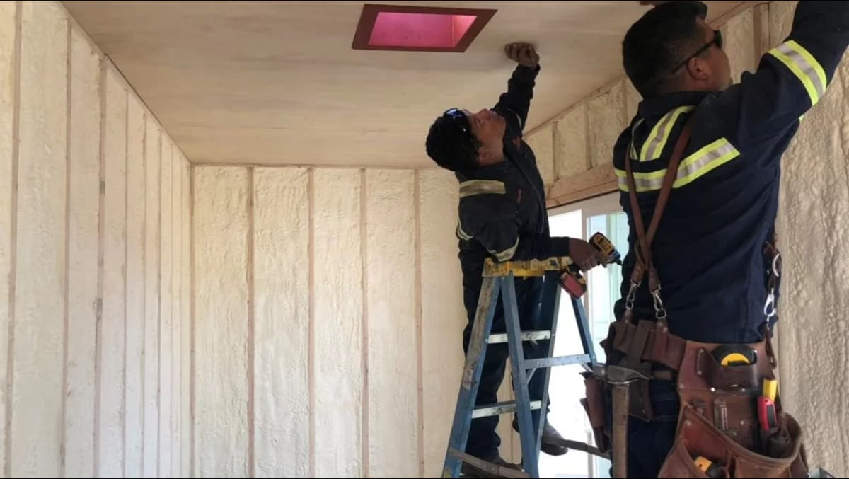 Two Conexwest technicians inside an insulated shipping container use tools and a ladder to install ceiling panels, with spray foam insulation visible on the walls.