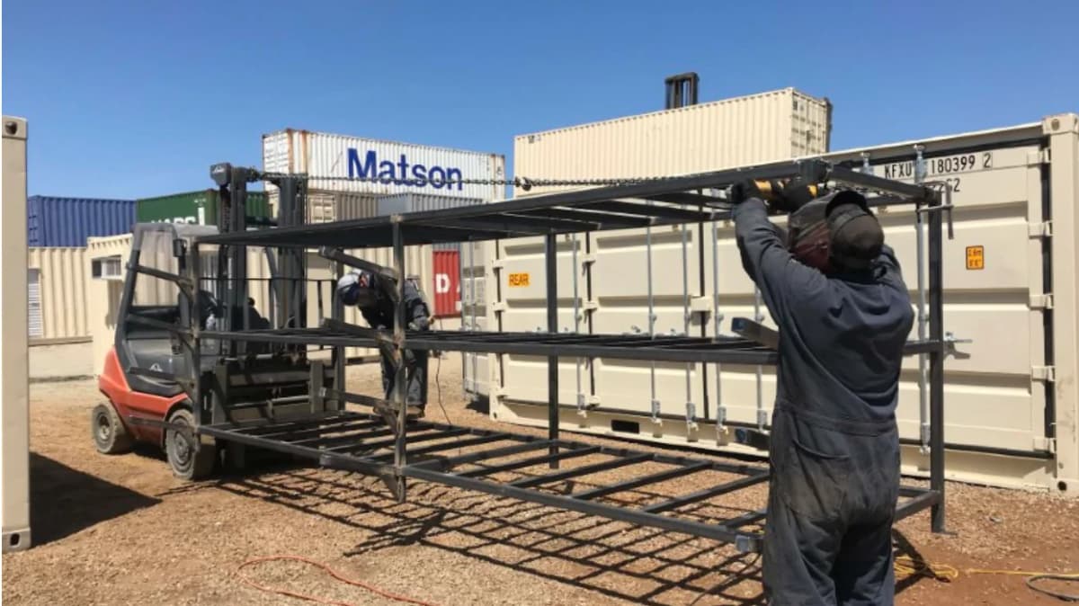 Two workers assembling a metal frame with a forklift near several stacked shipping containers