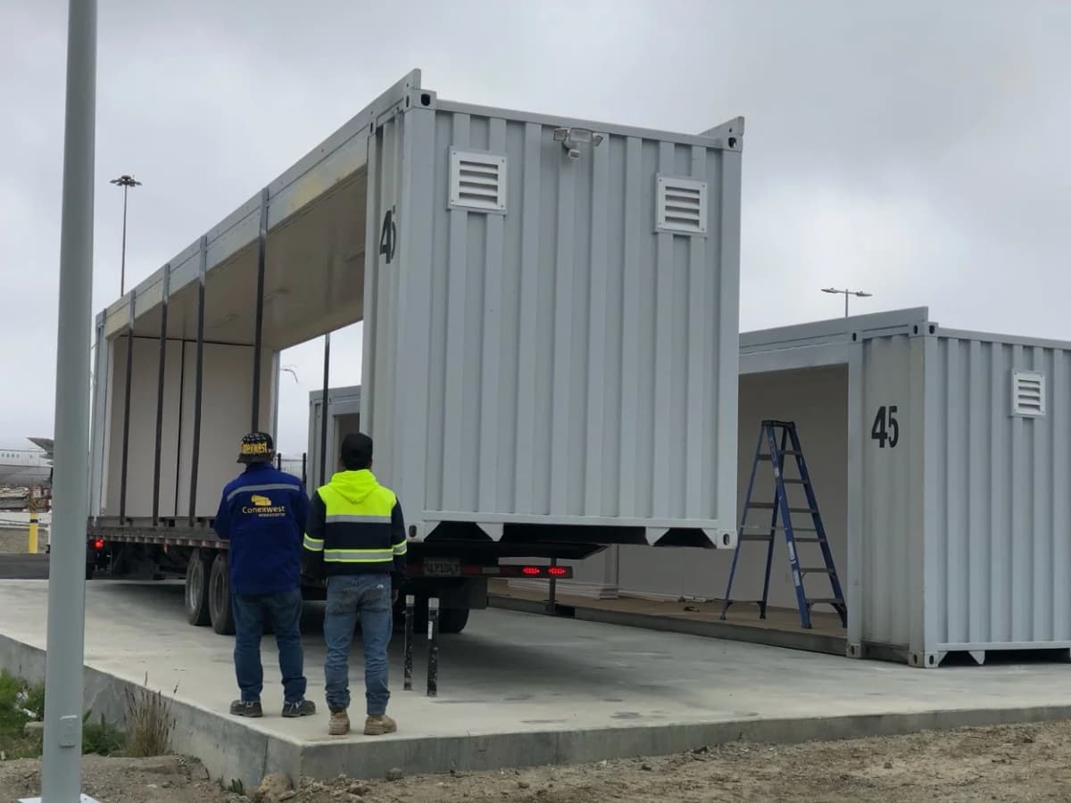 Two workers observing the installation of a modified shipping container with open sides at a construction site.
