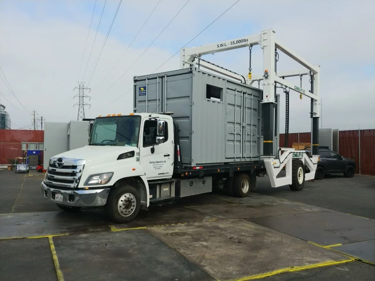 White commercial truck with hydraulic lifting crane apparatus positioned at an industrial container facility.