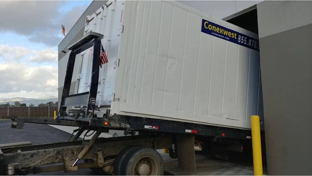 White shipping container being transported on a specialized truck trailer with hydraulic lift system.