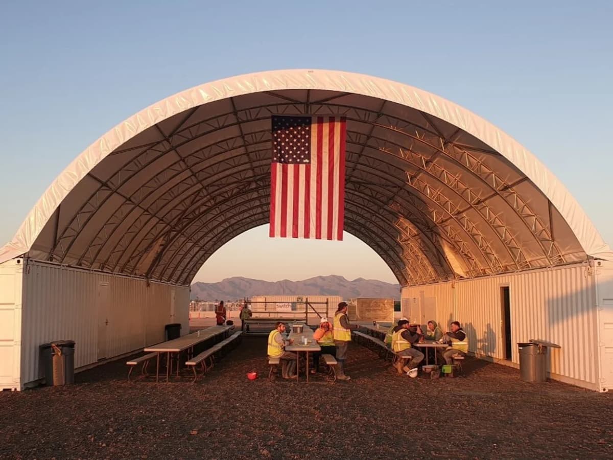 Workers sit and eat under a large canopy supported by two shipping containers