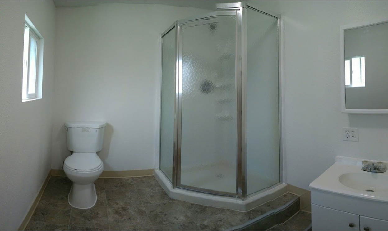Compact bathroom with a corner glass shower, white toilet, and small vanity sink, featuring neutral-colored flooring and minimal decor.