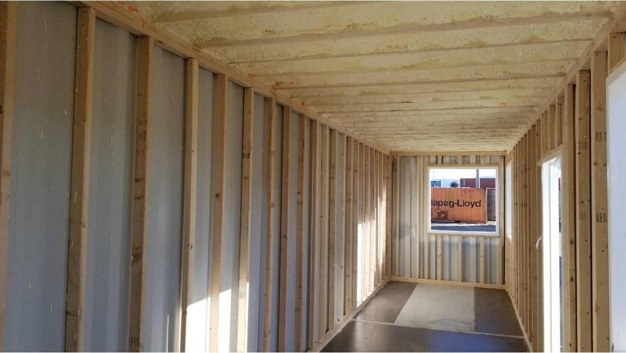 Interior construction view of a shipping container showing exposed corrugated steel walls, wooden wall studs, and ceiling beams.