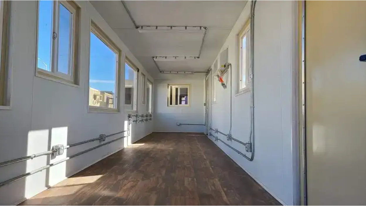 Interior corridor of a white container conversion with wood laminate flooring, multiple windows providing natural light, exposed wall fixtures, and track lighting on the ceiling.