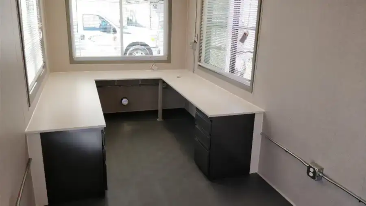 Interior of a compact container office with L-shaped white desks, black drawer units, and large windows on two sides.