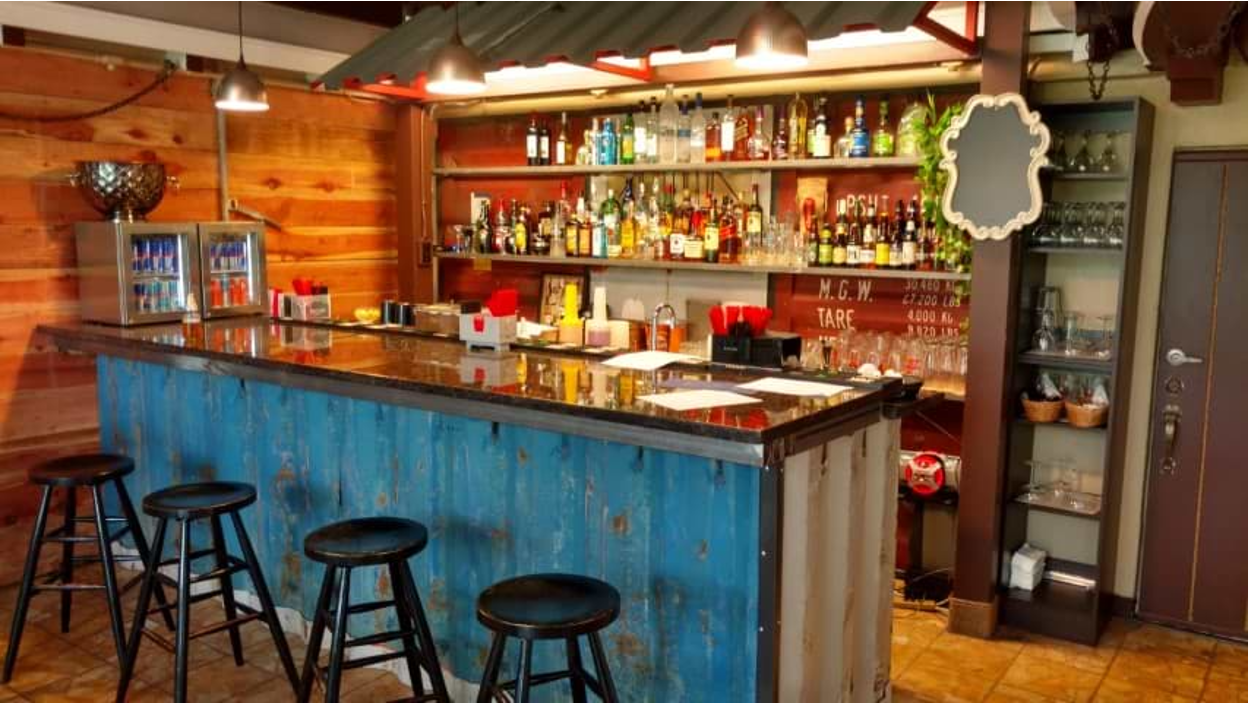 Interior view of a converted shipping container showing a fully equipped bar area with blue counter, bar stools, and extensive bottle collection on shelves.