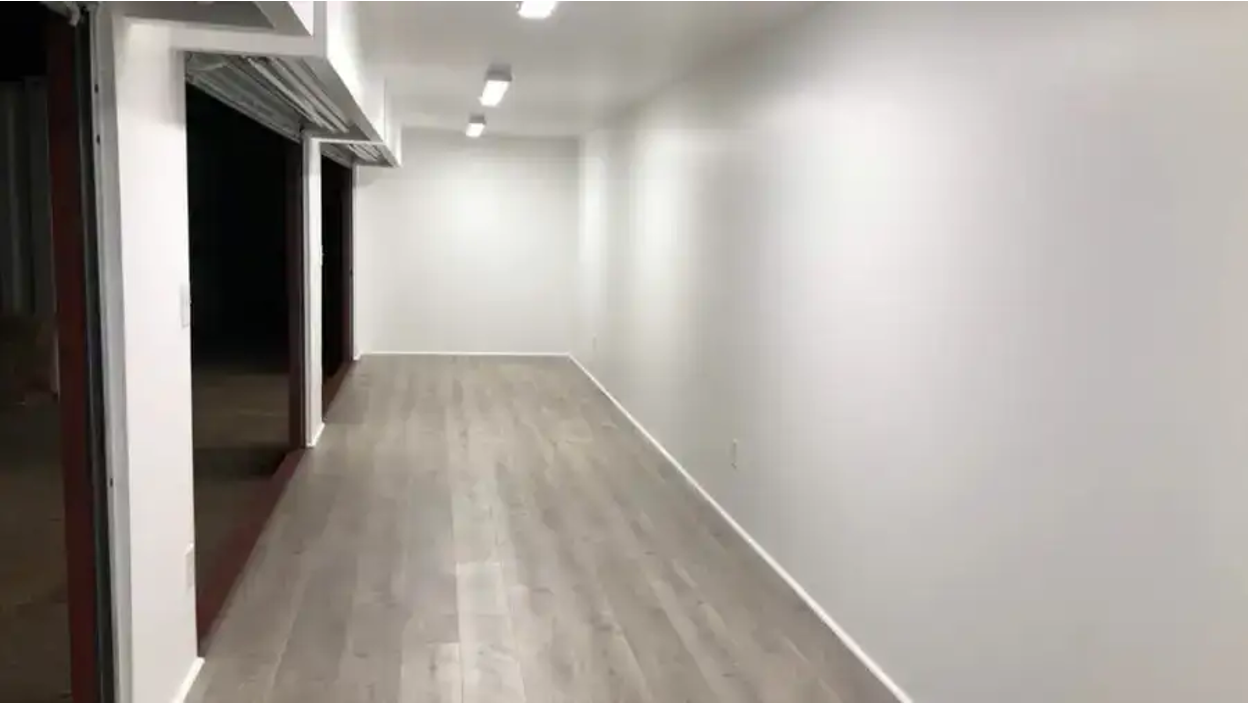Interior view of a renovated shipping container with white walls, ceiling lights, and light wood flooring.