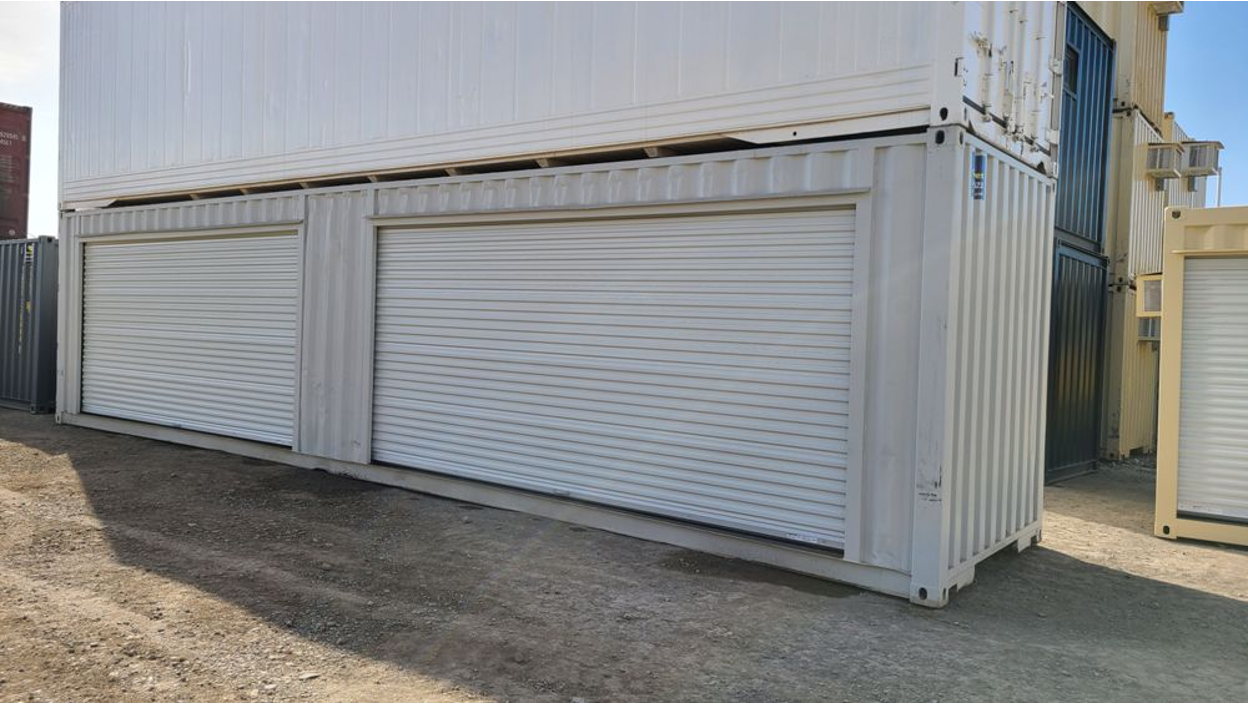 Stacked white shipping containers, with the lower container featuring two large white roll-up doors.