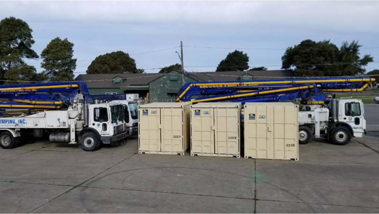 Three beige Conexwest storage containers are lined up in front of two large concrete pumping trucks with blue and yellow booms extended across the top.