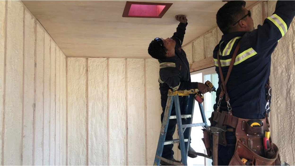 Two Conexwest technicians inside an insulated shipping container use tools and a ladder to install ceiling panels, with spray foam insulation visible on the walls.