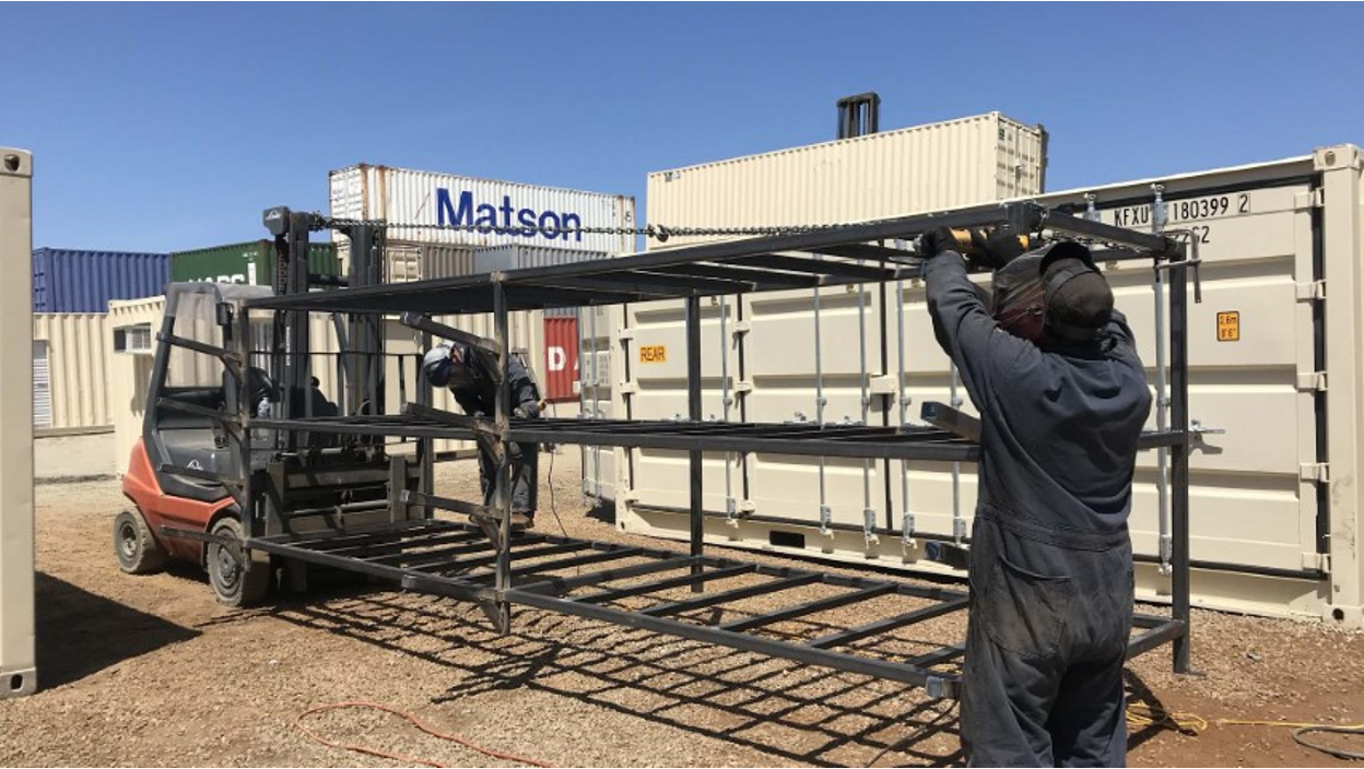 Two Conexwest workers assemble a metal frame with the help of a forklift in a container yard surrounded by stacked shipping containers.