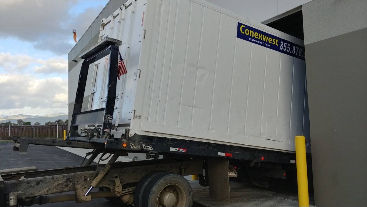 White shipping container being transported on a specialized truck trailer with hydraulic lift system.
